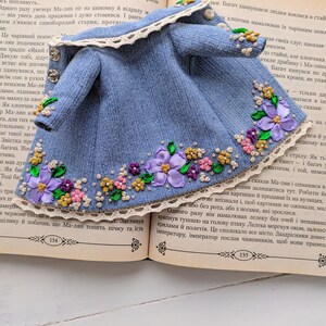 以下が含まれることがあります： 紫、ピンク、黄色の花柄刺繍が施されたミニチュアデニムジャケット。 クリーム色のレーストリムと小さなシルバーボタンが付いており、人形や小さなフィギュア向けにデザインされています。