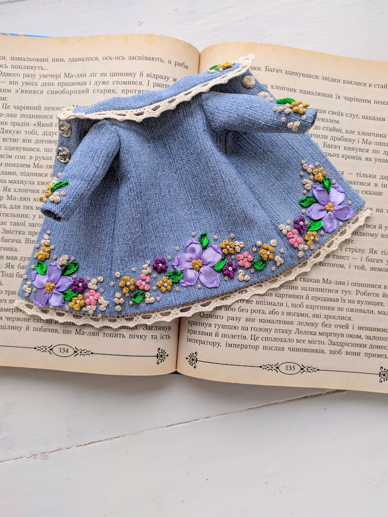 以下が含まれることがあります： 紫、ピンク、黄色の花柄刺繍が施されたミニチュアデニムジャケット。 クリーム色のレーストリムと小さなシルバーボタンが付いており、人形や小さなフィギュア向けにデザインされています。