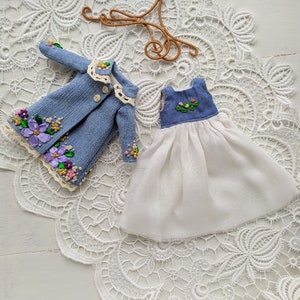 以下が含まれることがあります： 紫、ピンク、黄色の花の刺繍が施された、ミニチュアデニムジャケットとドレスのセット。 ジャケットはクリーム色のレースの襟とボタンが付いています。 ドレスは白いスカートと、花のアクセントが付いた青いボディスです。