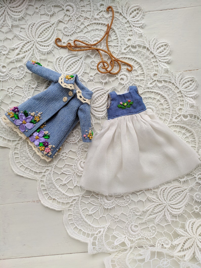 以下が含まれることがあります： 紫、ピンク、黄色の花の刺繍が施された、ミニチュアデニムジャケットとドレスのセット。 ジャケットはクリーム色のレースの襟とボタンが付いています。 ドレスは白いスカートと、花のアクセントが付いた青いボディスです。