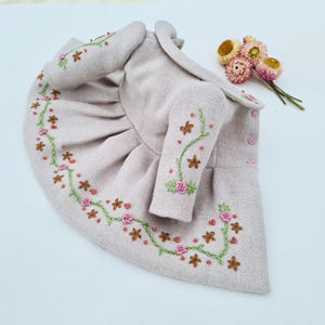 May include: A light pink coat with floral embroidery. The coat features a round collar, long sleeves, and a flared skirt. The embroidery includes pink roses, green vines, and brown star-shaped accents. A small bouquet of dried flowers is in the background.