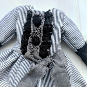 May include: A miniature dress with a black and white striped pattern. The bodice features black lace and rosettes, with a grey bow at the waist. The sleeves have black lace cuffs. A vintage-inspired garment.