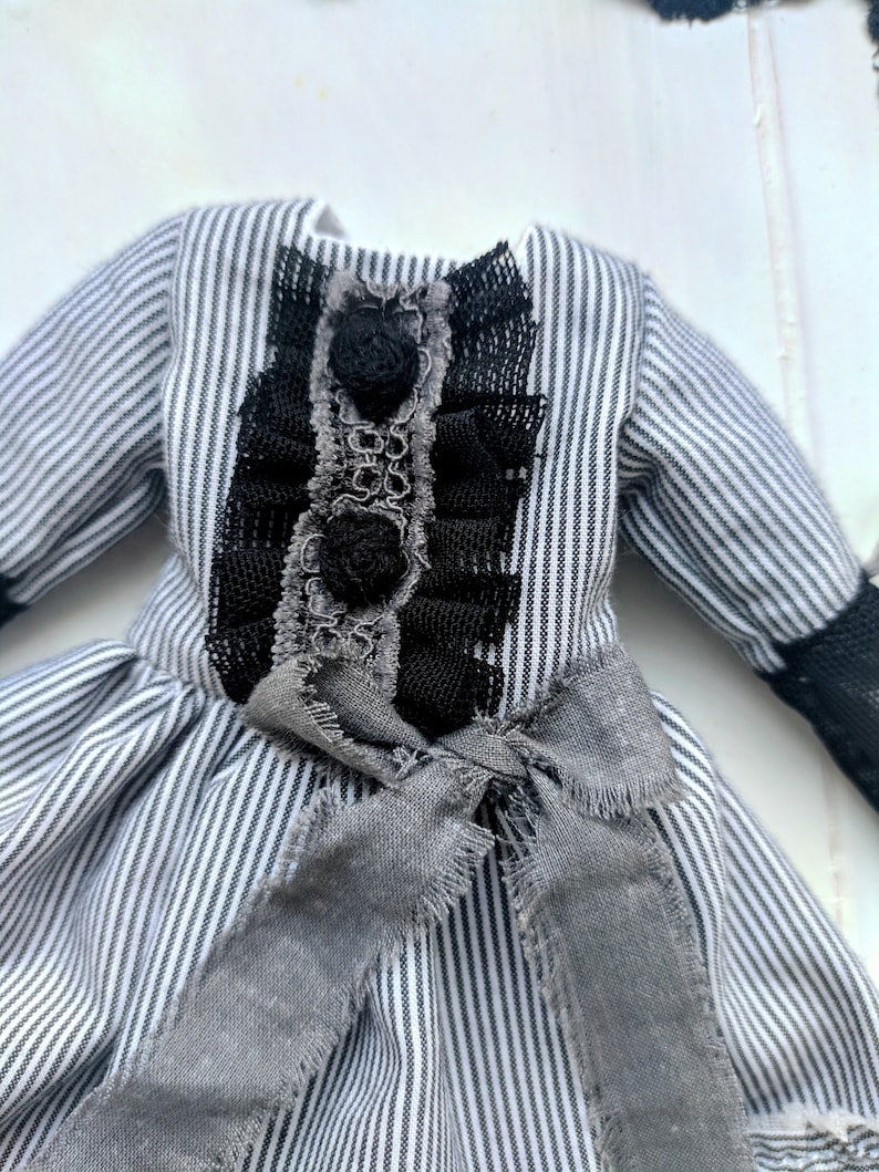May include: A miniature dress with a black and white striped pattern. The bodice features black lace and rosettes, with a grey bow at the waist. The sleeves have black lace cuffs. A vintage-inspired garment.