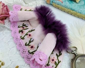 May include: A pink knitted jacket with a purple fur trim and floral embroidery. The jacket has a lace trim and is displayed on a white lace background.
