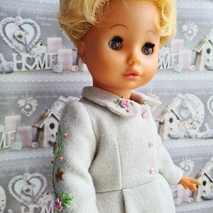 K&ouml;nnte beinhalten: Eine blonde Puppe, die ein graues Kleid mit Blumenstickerei tr&auml;gt. Das Kleid hat einen wei&szlig;en Kragen und rosa Kn&ouml;pfe. Die Puppe hat braune Augen und schaut nach rechts.