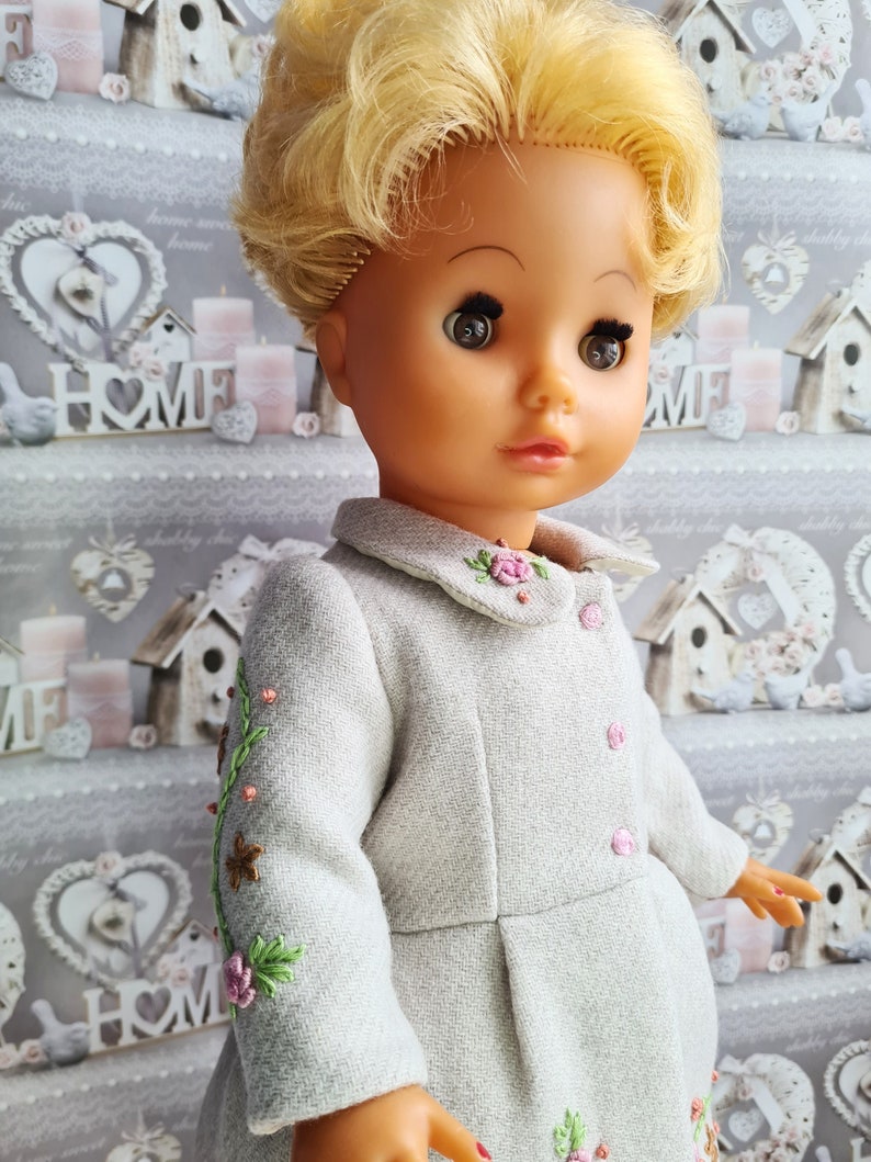 K&ouml;nnte beinhalten: Eine blonde Puppe, die ein graues Kleid mit Blumenstickerei tr&auml;gt. Das Kleid hat einen wei&szlig;en Kragen und rosa Kn&ouml;pfe. Die Puppe hat braune Augen und schaut nach rechts.