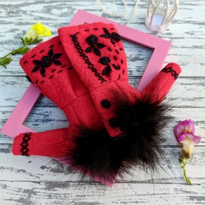 May include: A red felt doll coat with black embroidery and a black fur trim. The coat is lying on a white wooden surface with a pink picture frame and a white birdcage.