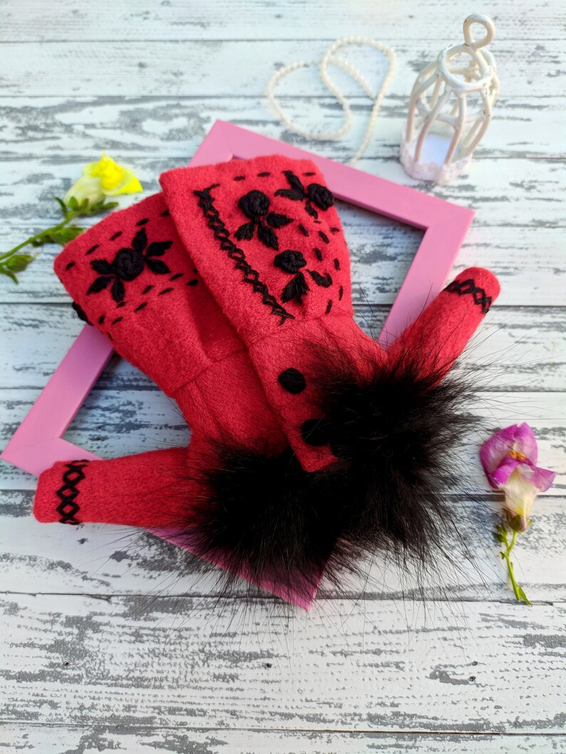 May include: A red felt doll coat with black embroidery and a black fur trim. The coat is lying on a white wooden surface with a pink picture frame and a white birdcage.