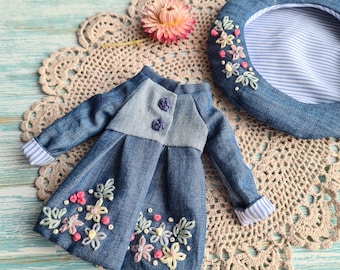 Puede incluir: Un conjunto de muñeca de mezclilla con chaqueta y boina. La chaqueta tiene un corpiño azul claro y una falda azul oscuro, adornada con flores bordadas en rosa, amarillo y blanco. La boina es de mezclilla con interior a rayas y bordados florales a juego.