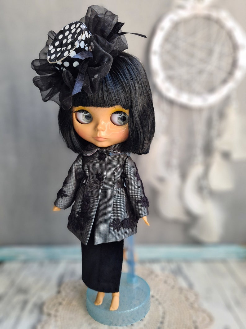 May include: A black and white polka dot hat with a black bow and veil on a doll with black hair, wearing a gray and black jacket and a black skirt. The doll is standing on a blue stand.