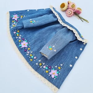 May include: A denim dress with long sleeves, adorned with embroidered flowers in shades of purple, yellow, and pink. The dress features a cream-colored lace trim along the edges and around the collar. The dress is designed for a child.