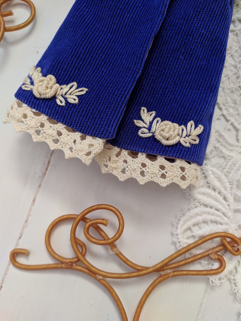 May include: Two blue fabric sleeves with white lace trim and white embroidered floral designs.