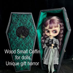 May include: A small wooden coffin with a green velvet lining and black lace trim. The coffin is open and a doll is inside. The doll has purple hair, a black and white dress, and a green sash. The text "Wood Small Coffin for dolls, Unique gift horror" is printed on the front of the coffin.