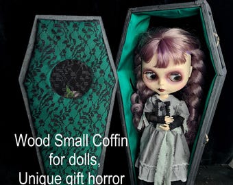 May include: A small wooden coffin with a green velvet lining and black lace trim. The coffin is open and a doll is inside. The doll has purple hair, a black and white dress, and a green sash. The text "Wood Small Coffin for dolls, Unique gift horror" is printed on the front of the coffin.