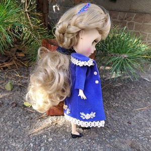 May include: A doll with long blonde hair styled in a braid, wearing a blue corduroy coat with white lace trim and floral embroidery. The doll is wearing black shoes and has a pink and blue hair clip.