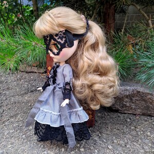 May include: A doll with blonde hair wearing a black lace eye mask and a grey and white striped dress with black lace trim. The doll has a grey sash tied around the waist and is standing on a grey surface.