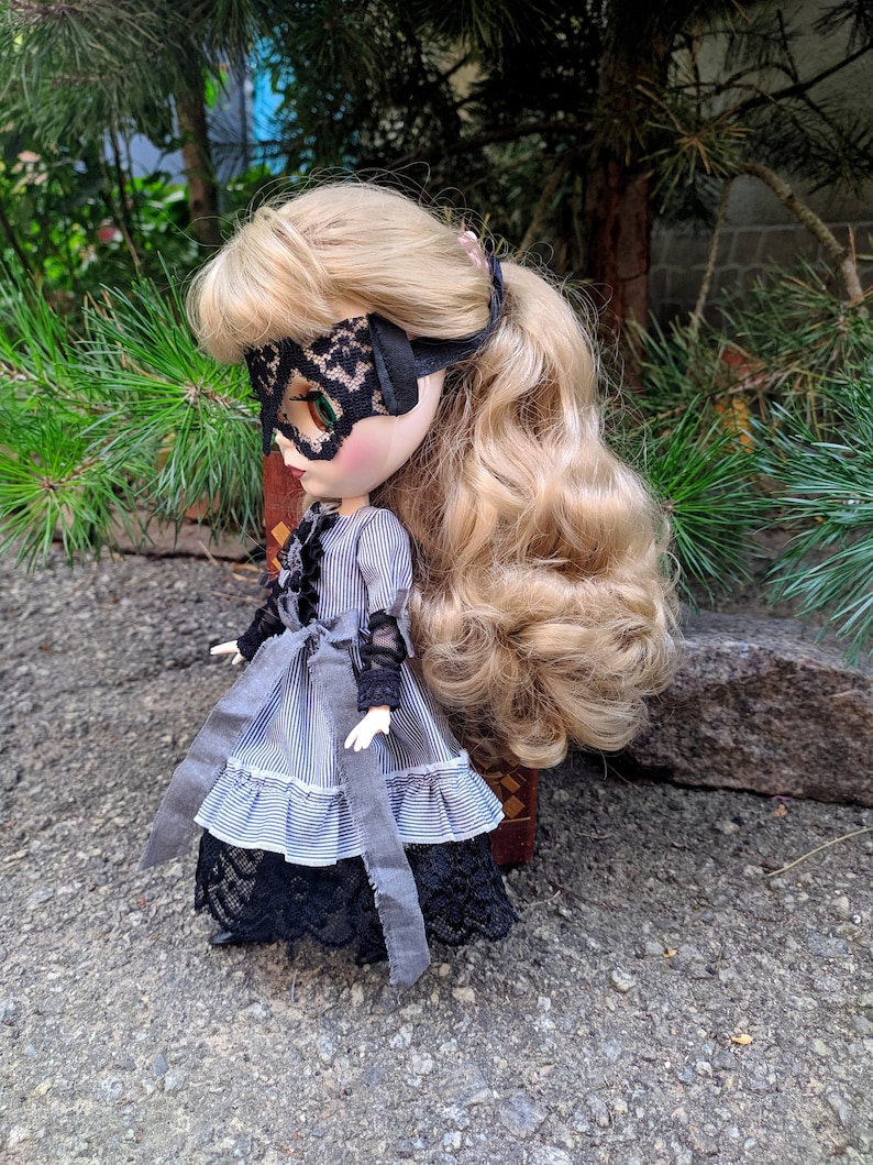 May include: A doll with blonde hair wearing a black lace eye mask and a grey and white striped dress with black lace trim. The doll has a grey sash tied around the waist and is standing on a grey surface.