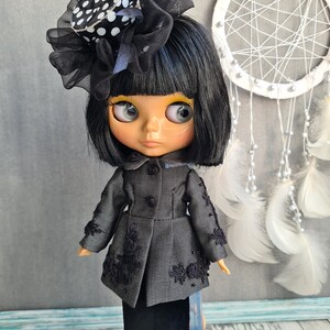 May include: A black and white polka dot hat with a black tulle bow sits atop a black wig on a doll with black eyes and yellow eyeshadow. The doll is wearing a black and white tweed jacket with black lace trim and a black skirt. The doll is standing on a blue stand.