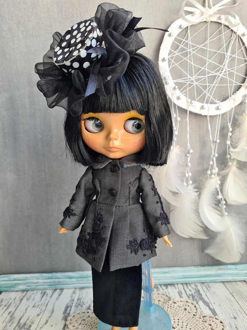 May include: A black and white polka dot hat with a black tulle bow sits atop a black wig on a doll with black eyes and yellow eyeshadow. The doll is wearing a black and white tweed jacket with black lace trim and a black skirt. The doll is standing on a blue stand.