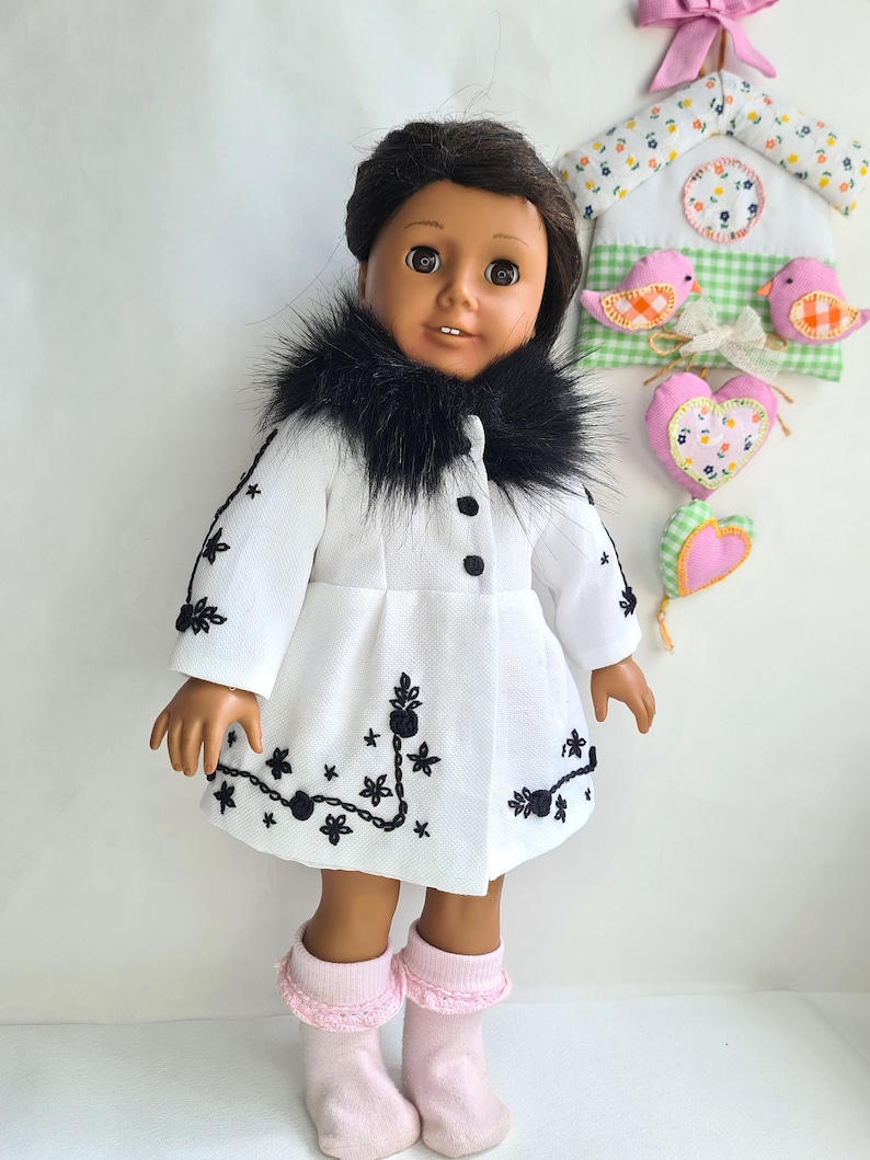 May include: A doll wearing a white coat with black embroidery and a black fur collar. The doll has brown skin, brown eyes, and black hair. The doll is wearing pink socks.