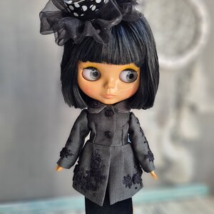 May include: A black and white polka dot hat with a black veil sits atop a doll's head. The doll has black hair, large eyes, and is wearing a black and white patterned coat and a black skirt.