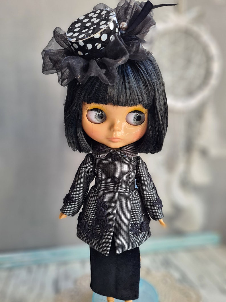 May include: A black and white polka dot hat with a black veil sits atop a doll's head. The doll has black hair, large eyes, and is wearing a black and white patterned coat and a black skirt.