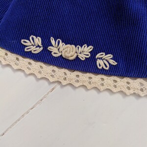 May include: A close-up of a blue fabric with white embroidered flowers and leaves. The fabric is trimmed with white lace.
