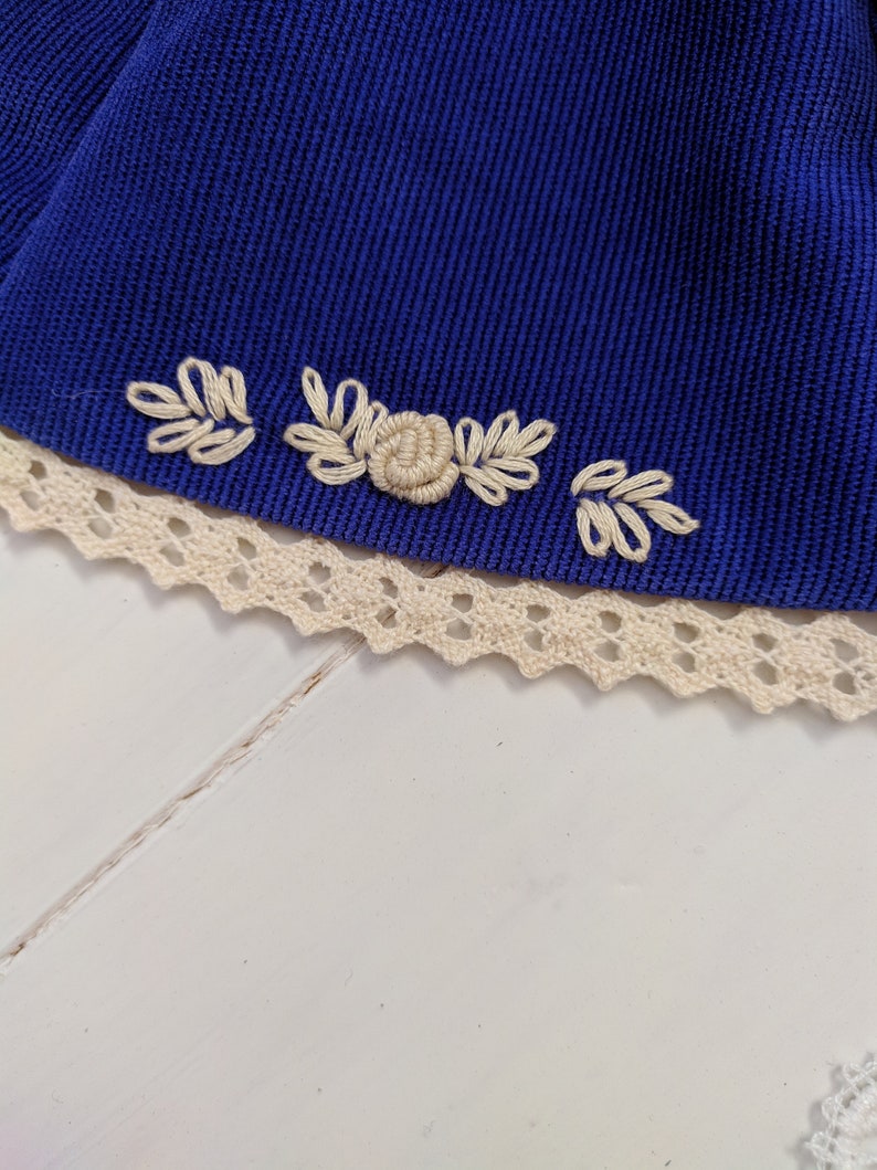 May include: A close-up of a blue fabric with white embroidered flowers and leaves. The fabric is trimmed with white lace.