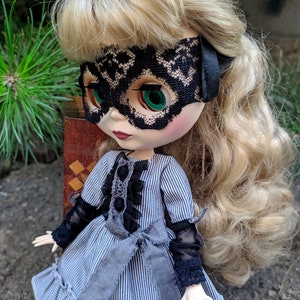 May include: A doll with blonde hair and green eyes wearing a black lace mask and a grey and white striped dress. The dress has black lace sleeves and a grey ribbon bow at the waist. The doll is posed outdoors.
