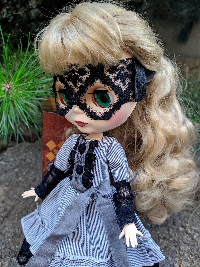 May include: A doll with blonde hair and green eyes wearing a black lace mask and a grey and white striped dress. The dress has black lace sleeves and a grey ribbon bow at the waist. The doll is posed outdoors.