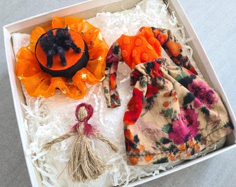 May include: A white box contains a Halloween-themed ensemble. It includes an orange and black spider decoration, a doll-like figure, and a floral-patterned outfit with an orange top. The ensemble is nestled in white paper.