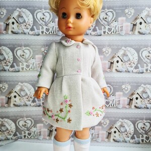 K&ouml;nnte beinhalten: Eine Puppe tr&auml;gt einen hellgrauen Mantel mit gestickten Blumen und wei&szlig;en Socken. Die Puppe hat blonde Haare und blaue Augen.