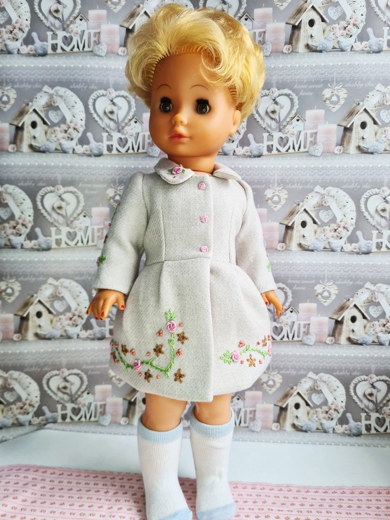 K&ouml;nnte beinhalten: Eine Puppe tr&auml;gt einen hellgrauen Mantel mit gestickten Blumen und wei&szlig;en Socken. Die Puppe hat blonde Haare und blaue Augen.