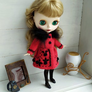 May include: A red felt doll coat with black embroidery and a black faux fur collar. The doll is wearing black tights and black shoes.