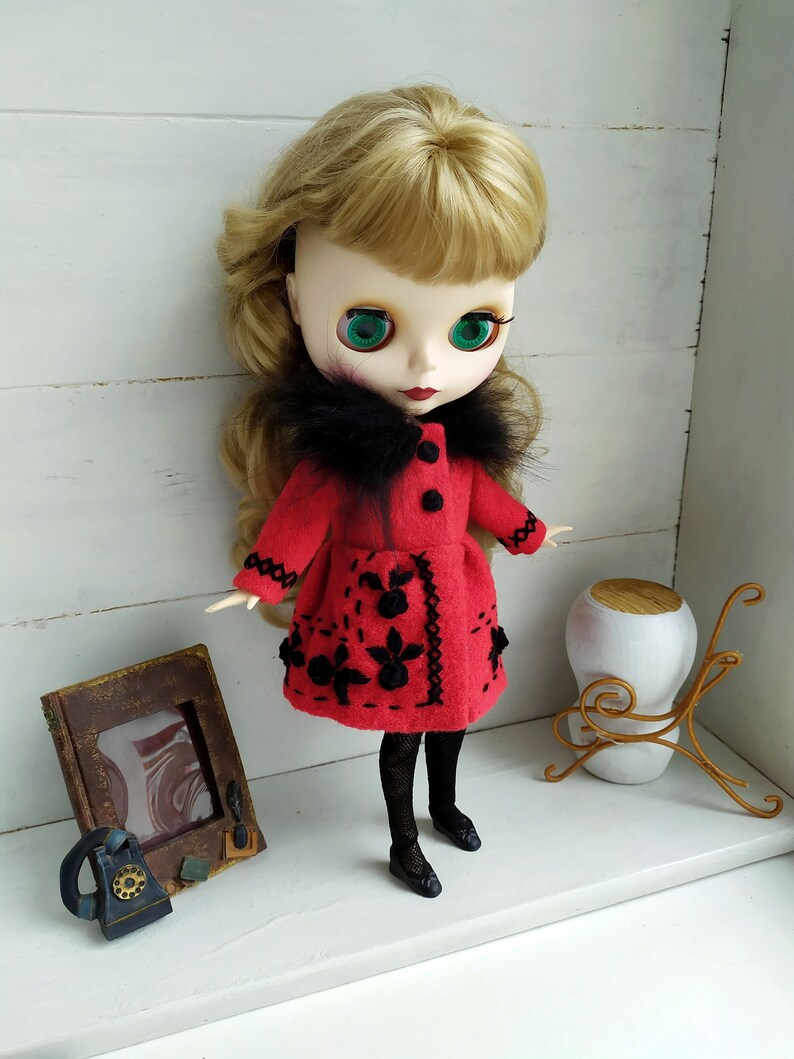 May include: A red felt doll coat with black embroidery and a black faux fur collar. The doll is wearing black tights and black shoes.