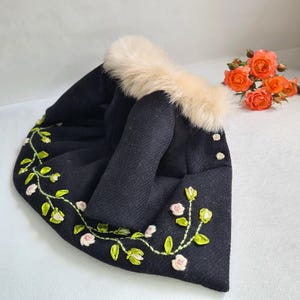 May include: A black hat with a cream-colored fur trim and floral embroidery. The hat features a decorative design of pink flowers and green leaves. Two small gold buttons are visible on the side.