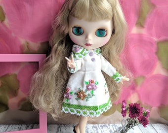 Puede incluir: Una muñeca Blythe con pelo largo rubio y grandes ojos verdes, vestida con un vestido blanco con bordados florales y ribete verde. La muñeca posa con un signo de paz, con un marco rosa y flores en el fondo.
