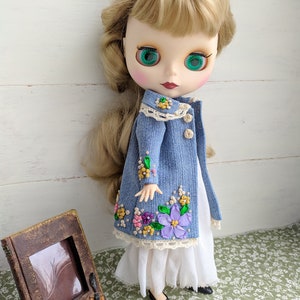 以下が含まれることがあります： 金髪と緑色の目をした人形。花柄の刺繍が入ったデニムジャケットと白いドレスを着ています。人形は黒い靴も履いています。ヴィンテージスタイルのフォトフレームと電話が背景にあります。