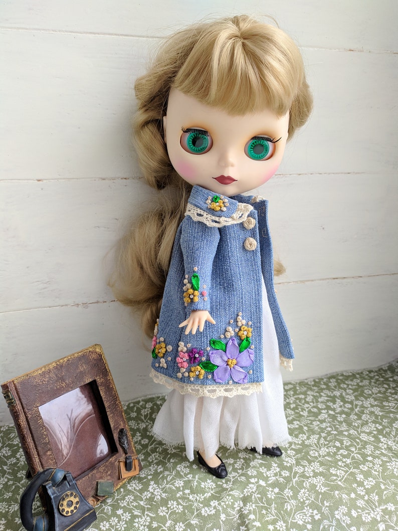 以下が含まれることがあります： 金髪と緑色の目をした人形。花柄の刺繍が入ったデニムジャケットと白いドレスを着ています。人形は黒い靴も履いています。ヴィンテージスタイルのフォトフレームと電話が背景にあります。