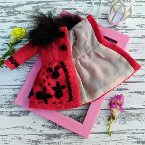 May include: A red felt doll coat with black embroidery and a black faux fur collar. The coat is open to reveal a gray lining. The coat is displayed on a pink frame against a white wooden background.