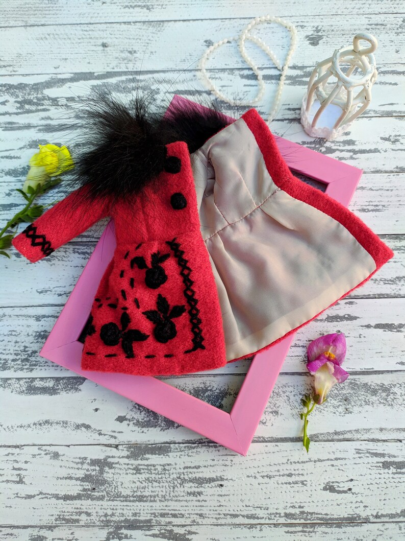 May include: A red felt doll coat with black embroidery and a black faux fur collar. The coat is open to reveal a gray lining. The coat is displayed on a pink frame against a white wooden background.