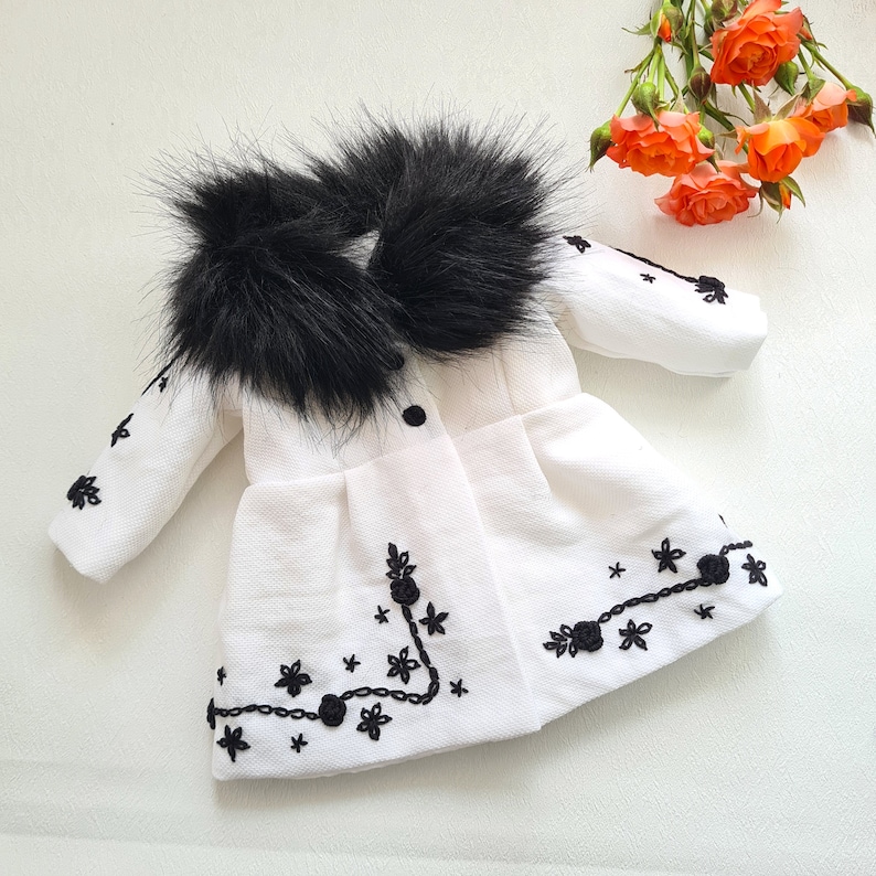 May include: A white faux fur coat with a black fur collar and black embroidered floral details.