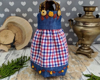 以下が含まれることがあります： デニムのボディスに刺繍の花と木製のボタンが付いた、ドレスのような布製のボトルカバー。スカートは赤と白のチェック柄。ハンドルは茶色です。
