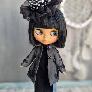 May include: A black and white polka dot hat with a black tulle bow sits on the head of a doll with black hair and a black dress. The doll has large, blue eyes and a light skin tone.