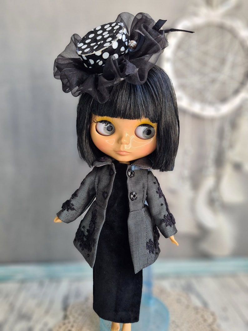 May include: A black and white polka dot hat with a black tulle bow sits on the head of a doll with black hair and a black dress. The doll has large, blue eyes and a light skin tone.