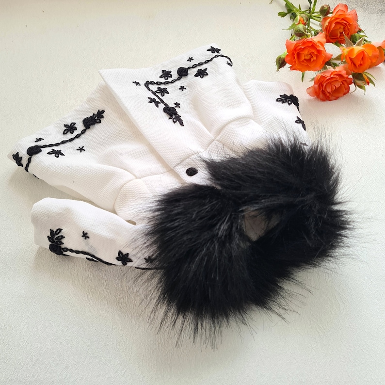 May include: White embroidered coat with a black faux fur collar. The coat has a button closure and is decorated with black embroidery.