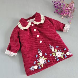 May include: A burgundy corduroy coat with a white lace collar and floral embroidery. The coat features white buttons and floral patterned cuffs. The embroidery includes pink, blue, and green floral designs, creating a charming, vintage aesthetic.
