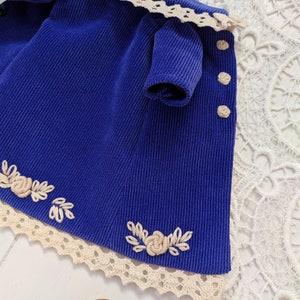May include: A blue corduroy dress with white lace trim and embroidered flowers. The dress has a small sleeve and buttons down the front.