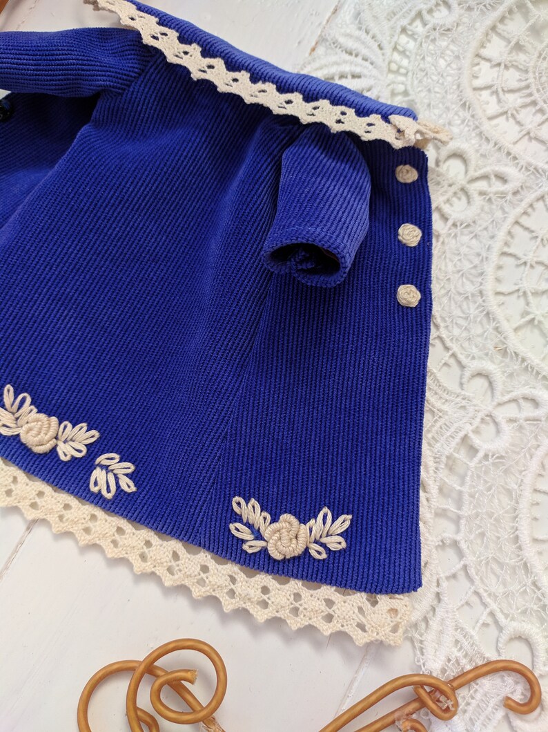 May include: A blue corduroy dress with white lace trim and embroidered flowers. The dress has a small sleeve and buttons down the front.