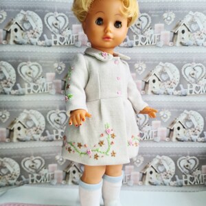 K&ouml;nnte beinhalten: Eine Vintage-Puppe mit blonden Haaren, die ein wei&szlig;es Kleid mit gestickten Blumen und blauen Socken tr&auml;gt. Die Puppe steht auf einer rosa-wei&szlig;en gemusterten Oberfl&auml;che.